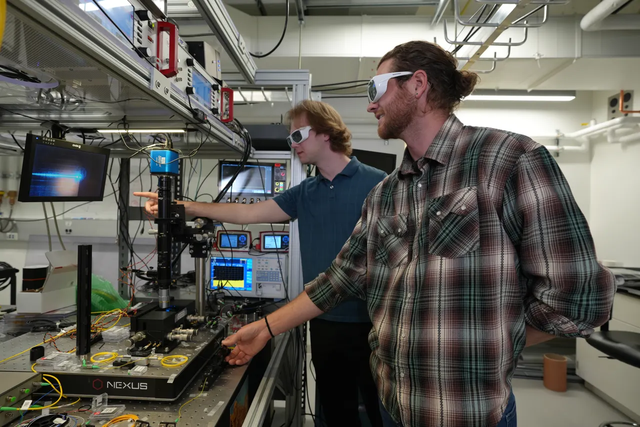 Two male researchers in casual clothes and protective eyewear stand in the lab, pointing at a screen hooked up to a laser science instrument.