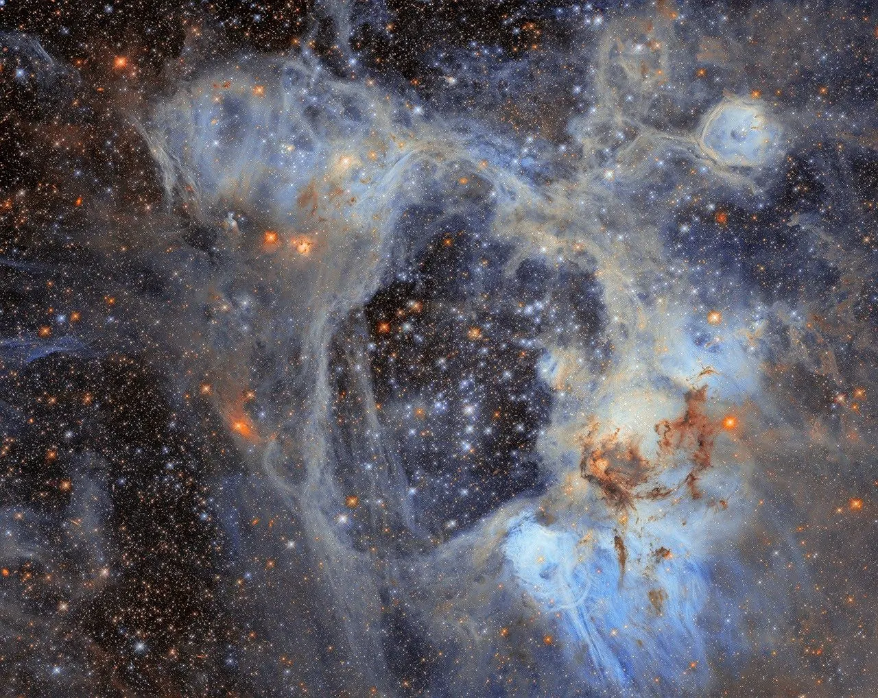 A dense field of stars fills the image, surrounded by wispy clouds of gas and dust. Pale blue and grey nebulosity forms an intricate web across the scene, with darker clouds of dust concentrated towar