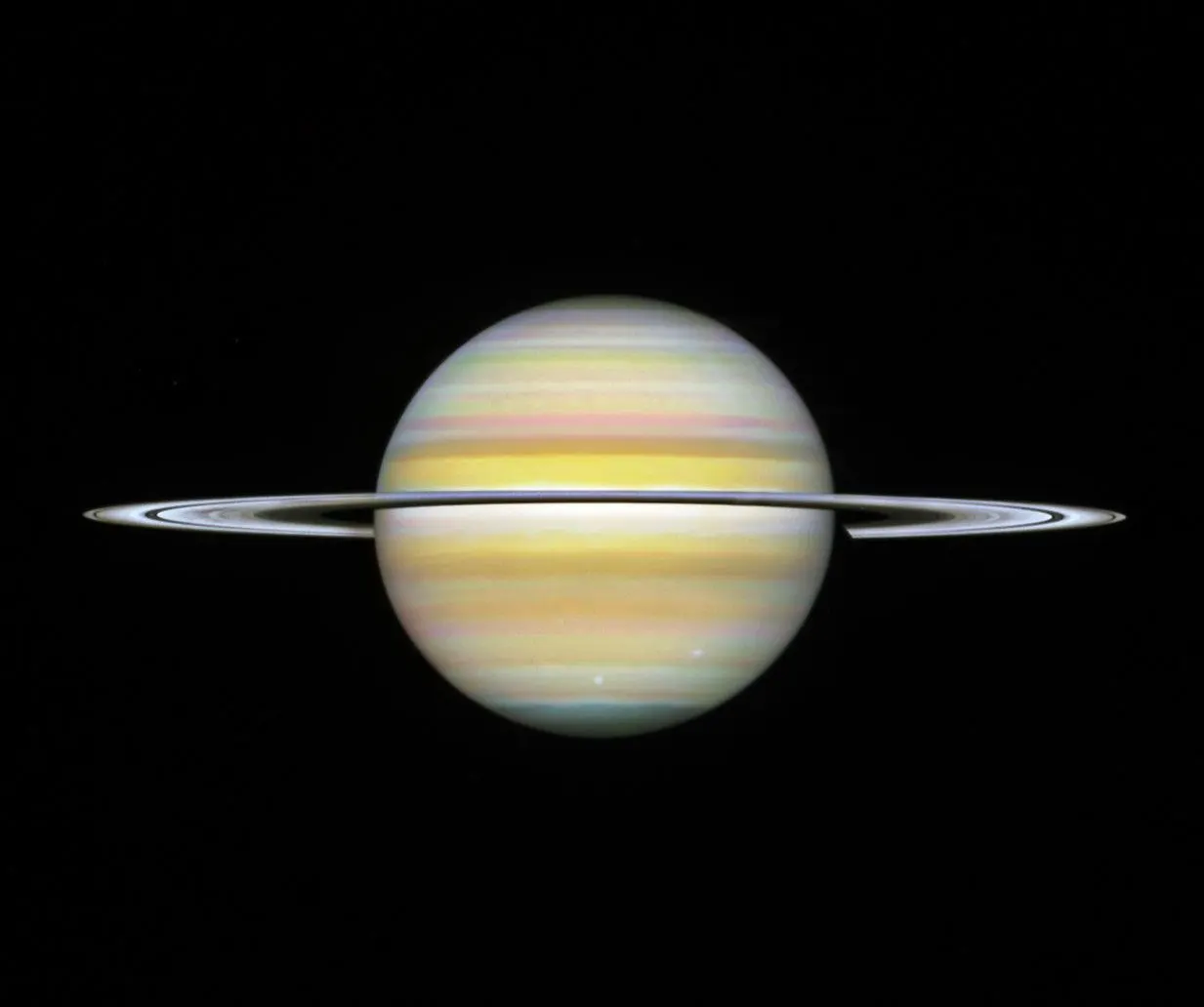 A straight-on view of Saturn, a globe with pale yellow horizontal bands at the equator and orange and pink at the mid-latitudes. Some bands towards the north and south pole have a light blue hue. Ther