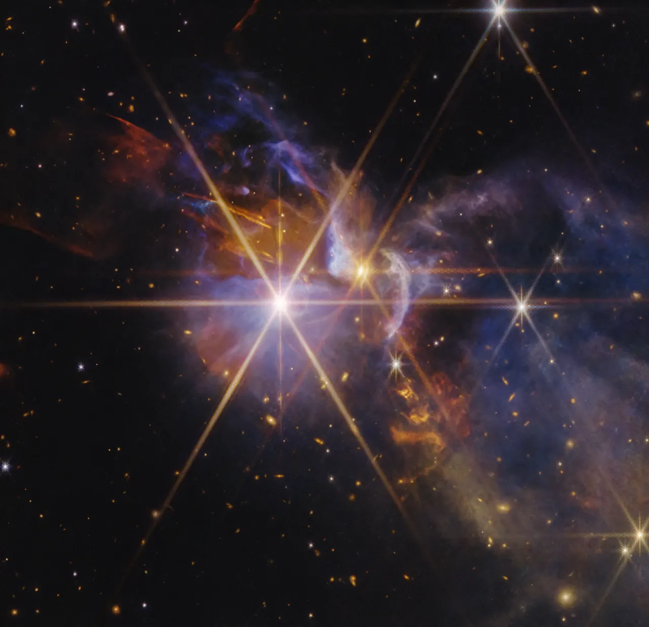 FS Tau, a star-forming nebula. Clouds of transparent blue and purple gas and dust stretch from the center to the right. Several yellow and white protostars, some showing Webb’s eight-pronged diffracti