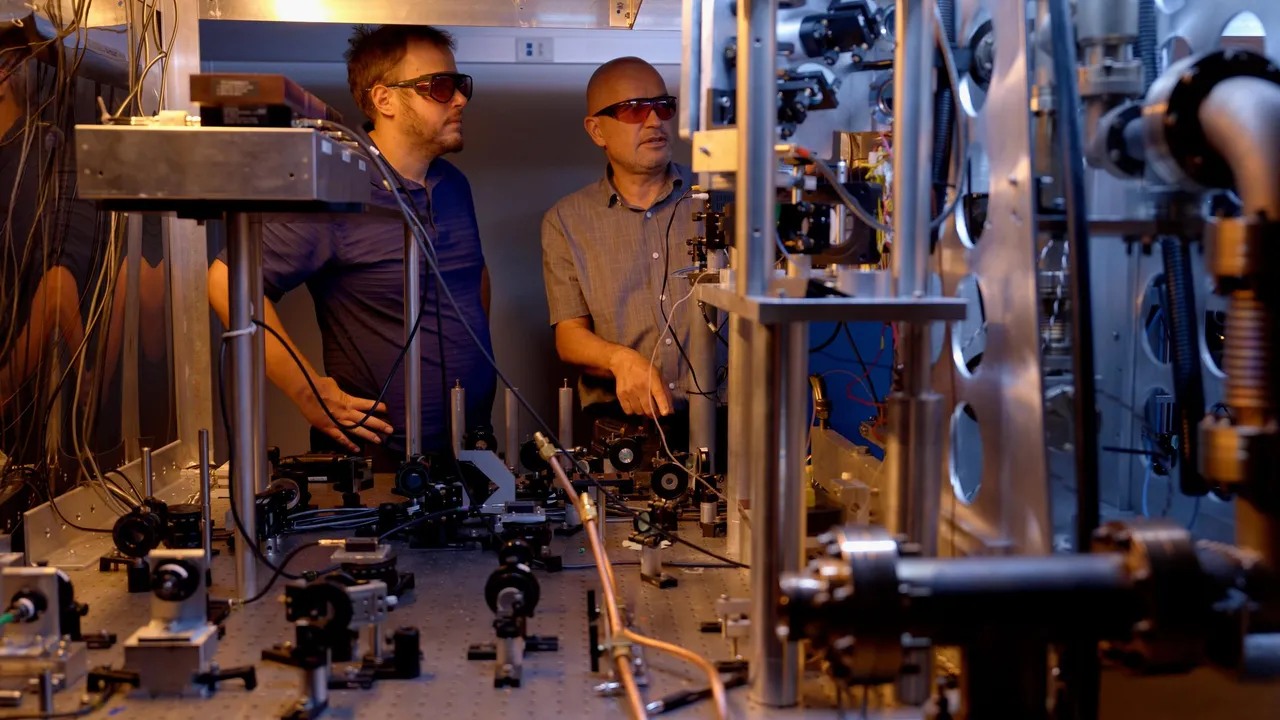 Two researchers in safety glasses stand behind a table holding various electronic and laser devices. A metal structure in the foreground is part of the atomic clock.
