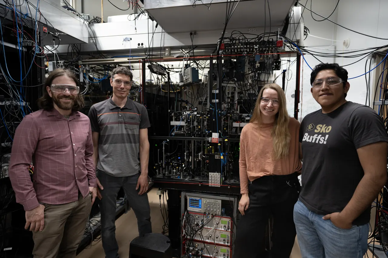 Four researchers pose standing around a large piece of scientific equipment covered with wiring and electronics.