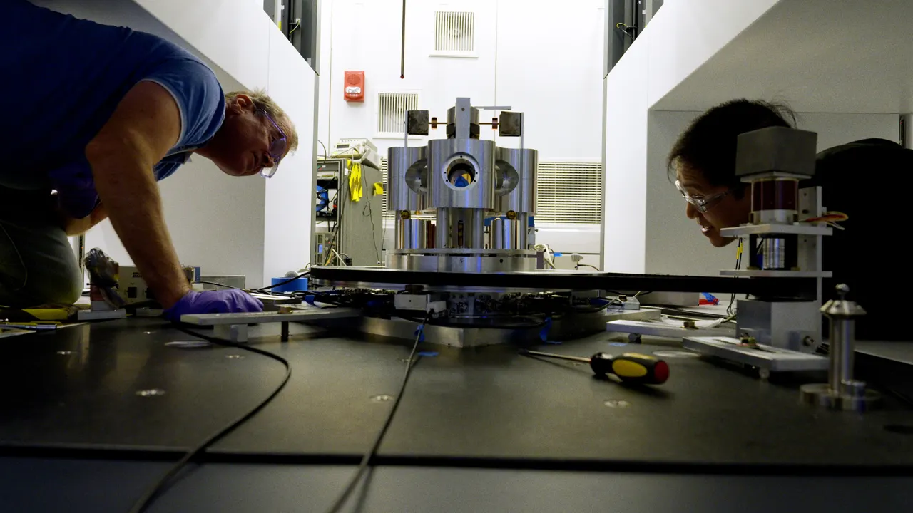 Wide shot shows two researchers on either side of a scientific device made of metal cylinders, peering closely at it.