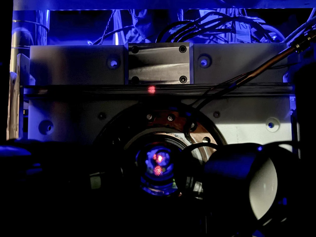 Close-up of a scientific instrument with wires and a circular metallic component, illuminated by blue light, featuring a small red dot at the center.